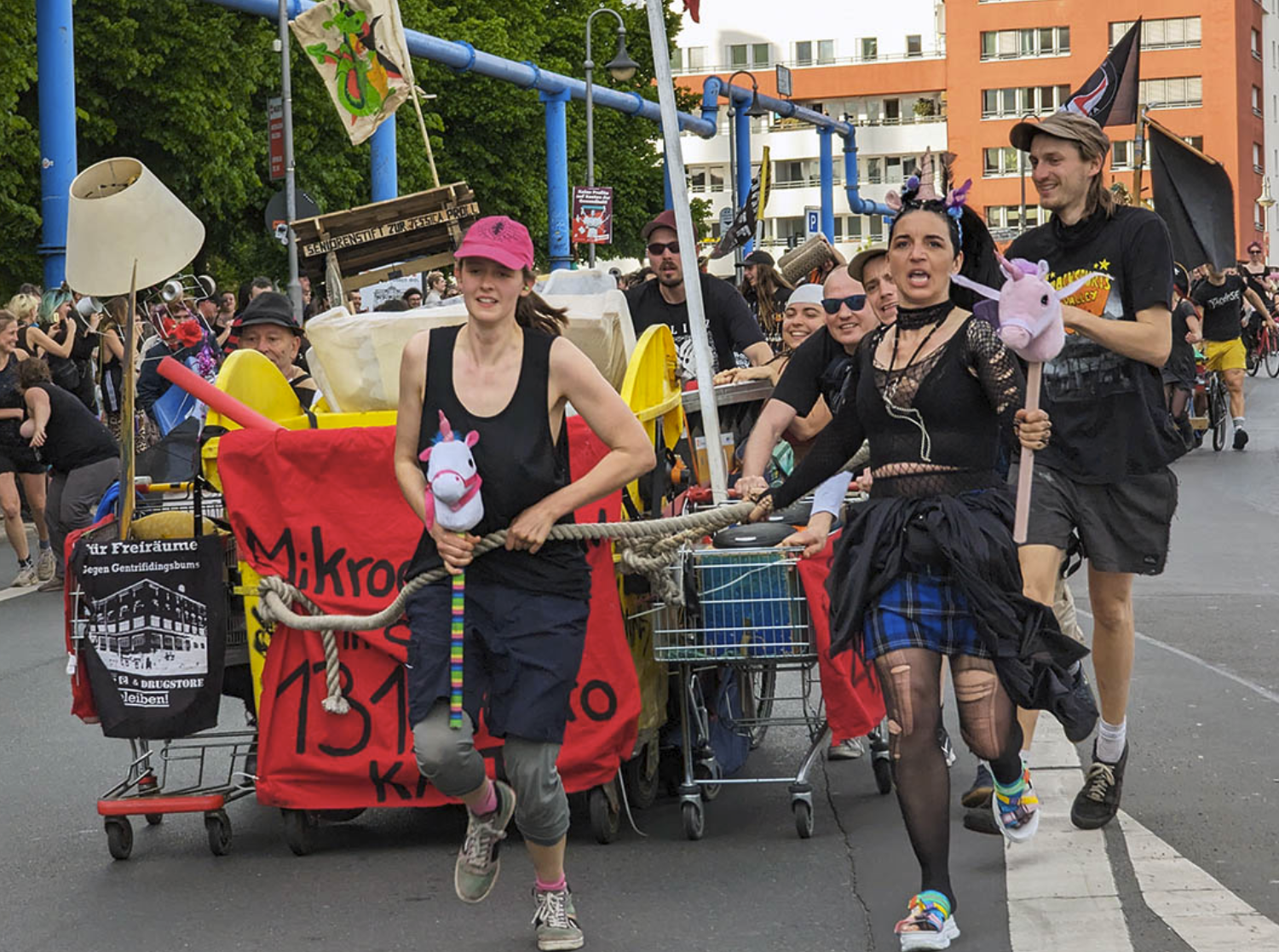 Das Foto zeigt einen Teil der Demo mit einem von mehreren Personen geschobenen aus Mülleimern und Einkaufswägen gebildeten Gefährt, an dem Transparente hängen.