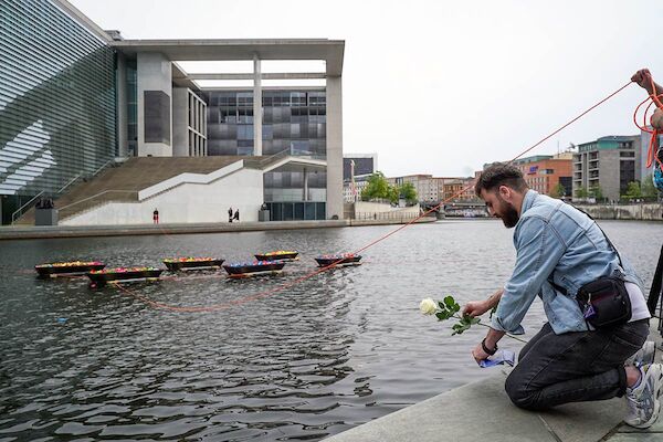 Das Foto zeigt einen Mann, der bei der Gedenkveranstaltung eine weiße Rose ins Wasser der Spree legt, in dem einige Sarguntereilte mit vielen bunten Papierschiffchen schwimmen.