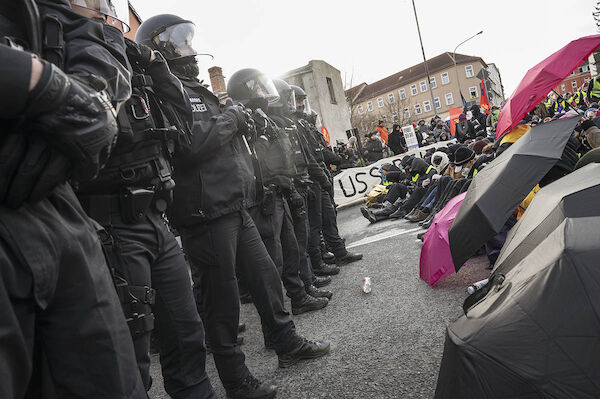 Fas Foto zeigt eine Sitzblockade, vor der sich zahlreiche martialische Polizisten in Kampfanzug aufgebaut haben.