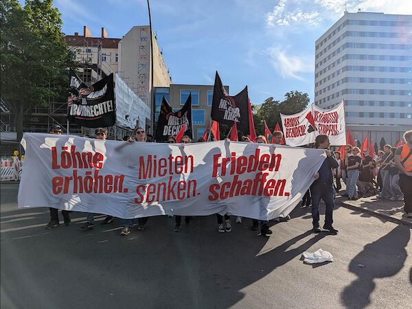 Das Foto von © Sabine Scheffer zeigt das Fronttransparent mit der Losung „Löhne erhöhen, Mieten senken, Frieden schaffen“ und dahinter laufende Menschen. Im HIntergrund sind weitere Transparente zu sehen.