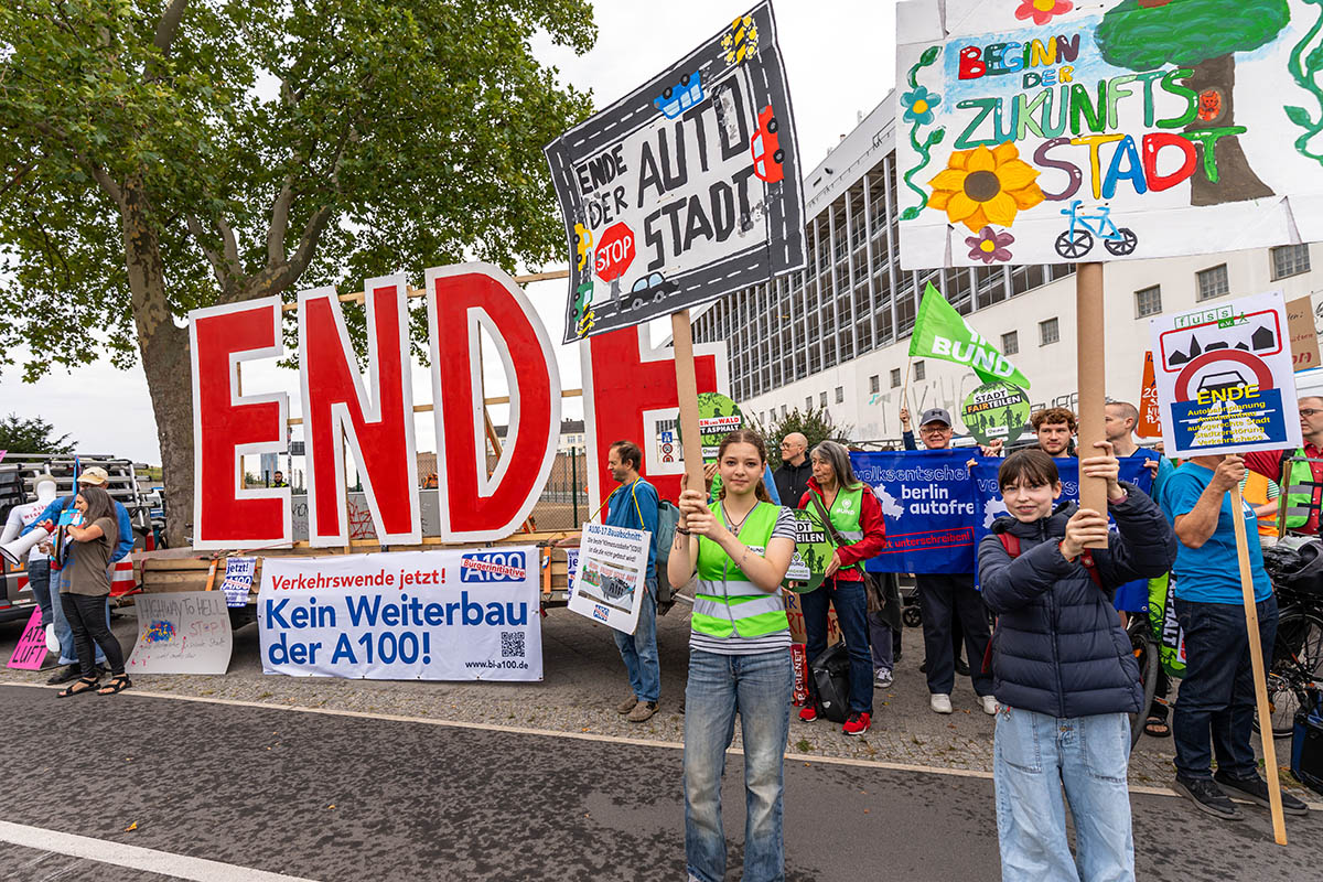 Das Foto zeigt einen kleinen Ausschnitt der Proteste mit einigen Menschen vor einem Anhänger, auf dem das Wort "Ende" in großen Buchstaben steht. Im Vordergrund stehen zwei junge Frauen, die gemalte Tafeln in die Höhe halten, die links mit dem Text "Ende der Autostadt" vor dem Hintergrund einer Asphalt- und Betonwüste, die rechts mit "Beginn der Zukunfstsstadt" und einem Baum und Blumen auf der Tafel.