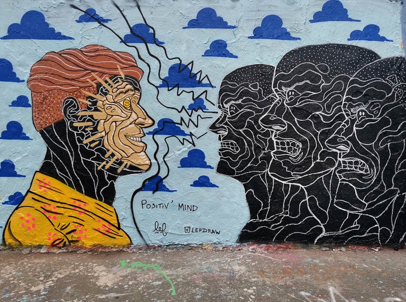Das Foto zeigt eine Wand mit einem Grafifti, das 3 Köpfe, die mit grimmiger Mine einen vierten anstarren, der freundlich zurück lächelt. Signiert ist das Bild mit dem Titel "Positiv' Mind" von Lefdraw (Instagram)