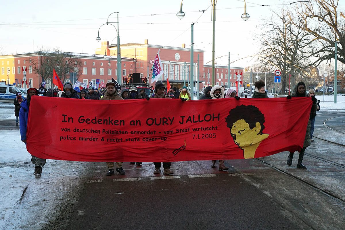 Start der Demo vor dem Bahnhof in Dessau mit dem Fronttransparent mit dem Text "In Gedenken an Oury Jalloh - von deutschen Polizisten ermordet, vom Staat vertuscht - police murder and racist state cover-up 7.1.2005" sowie ein Bild von Oury Jalloh.