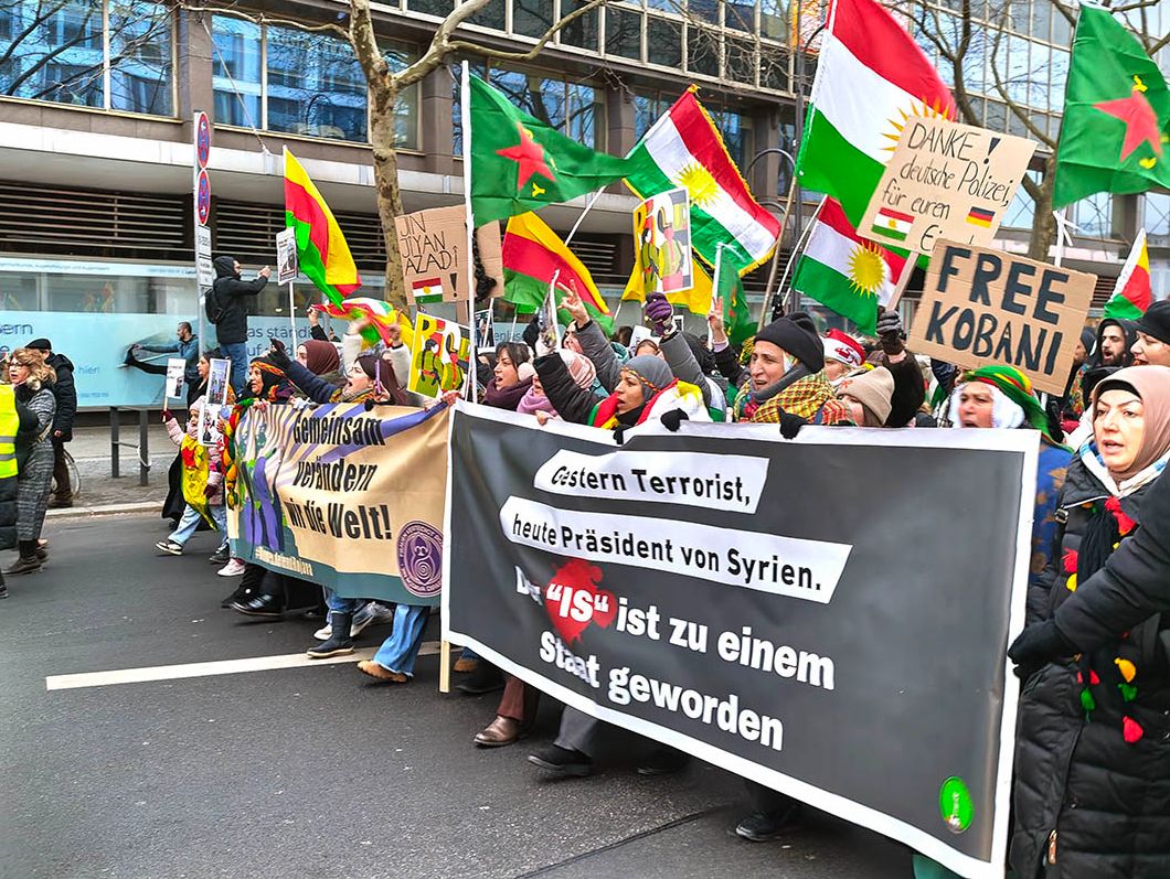 Das Foto von Sabine Scheffer zeigt einen Demoblock, der von rechts im Bild kommt und zwei nebeneinander getragene Fronttransparente mit dem Text Gestern Terrorist, heute Präsident von Syrien - Der "IS" ist zu einem Staat geworden" im Vordergrund und "Gemeinsam verändern wir die Welt" rechts davon , hinter denen Frauen laufen. Zum Teil werden YPG Fahnen getragen oder Papptafeln mit Texten wie "Free Kobani"