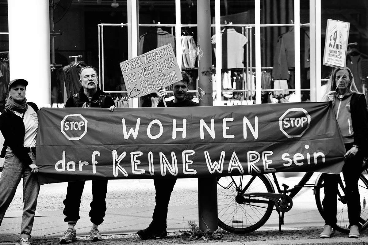 Das Foto von © neukoellnbild zeigt mehrere Personen an einem Transparent mit dem Text: "Stop - Wohnen darf keine Ware sein!" Eine Person trägt eine Papptafel mit dem Text "Unser Zuhause ist nicht Euer (Bild eines Scheißhaufens) Eigentum". Eine weitere Person trägt eine Plakattafel mit dem Mobiplakat zur Demo am 5.9.