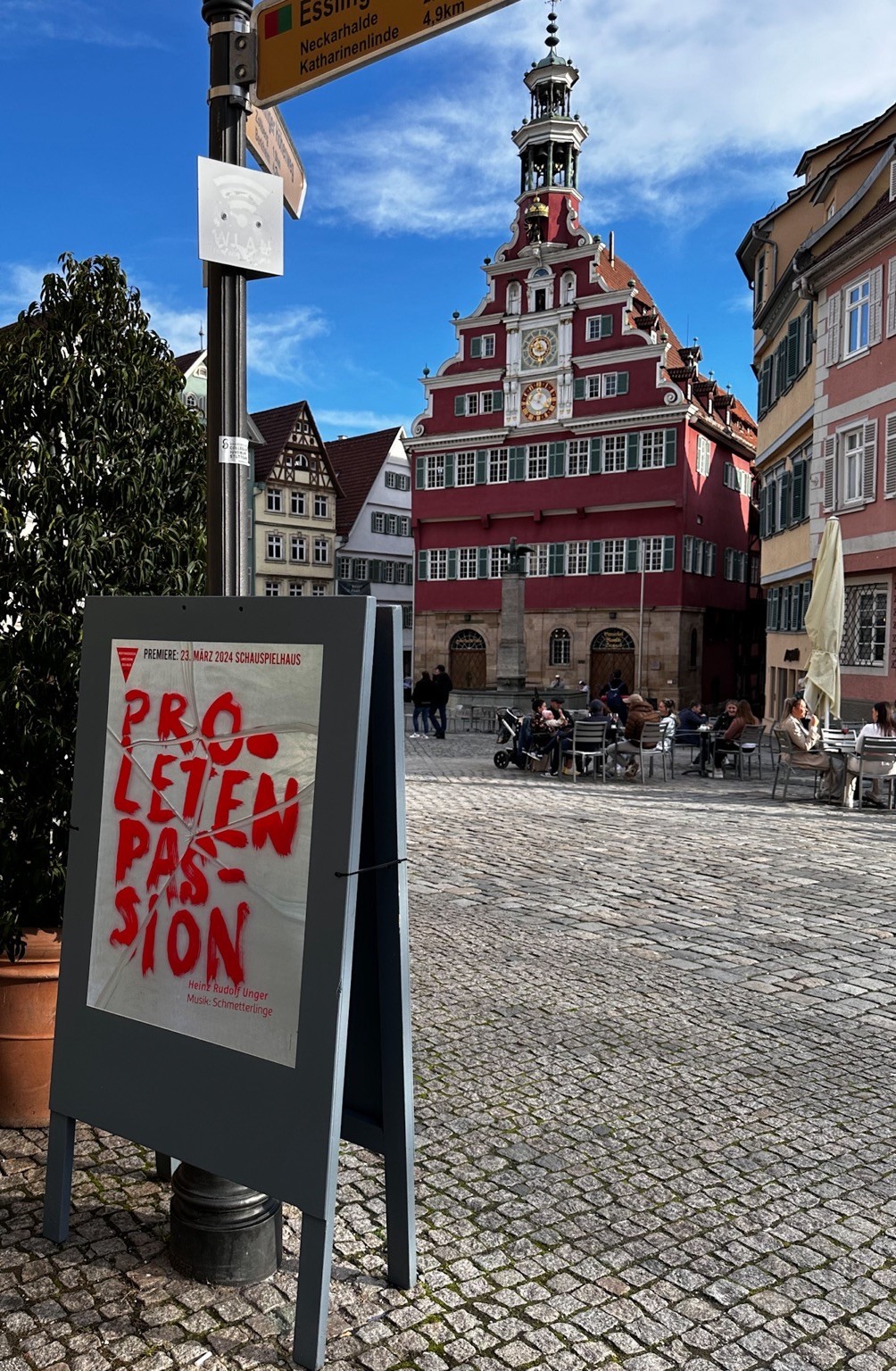 Foto: © Thomas Trueten Das Foto zeigt das Plakat der WLB zum Stück unter einem Wegweiser, im Hintergrund das alte Esslinger Rathaus, dazwischen sitzen Menschen in einem Straßencafe