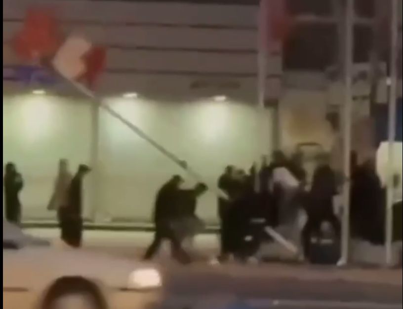 Menschen an einer Straße reißen einen Fahnenmast mit der iranischen Fahne um.