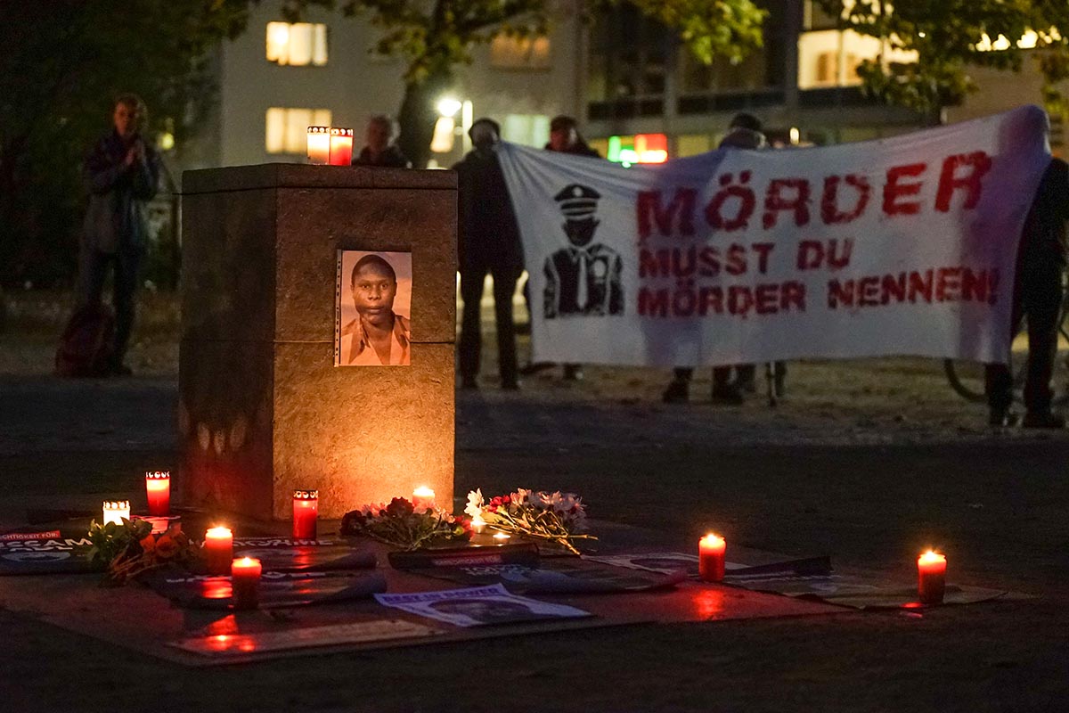 Das Foto zeigt ein Bild von Kupa Ilunga Medard Mutombo am Gedenkstein für die Opfer von Polizeigewalt, im Hintergrund Demonstrant:*Innen mit einem Transparent mit der Aufschrift "Mörder musst Du Mörder nennen!" Das Foto zeigt ein Bild von Kupa Ilunga Medard Mutombo am Gedenkstein für die Opfer von Polizeigewalt, im Hintergrund Demonstrant:*Innen mit einem Transparent mit der Aufschrift "Mörder musst Du Mörder nennen!"