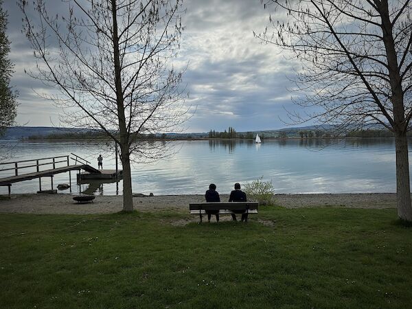Blick auf den Bodensee vom Naturfreundehaus aus: Zwei Personen sitzen auf einer Bank und blicken auf den See, auf dem ein Segelboot treibt. Links im Bild ist ein Bootssteg zu sehen, auf dem eine weitere Person steht. Regenwolken ziehen auf, im ruhigen See spiegeln sich die Bäume des gegenüber liegenden Ufers. Blick auf den Bodensee vom Naturfreundehaus aus: Zwei Personen sitzen auf einer Bank und blicken auf den See, auf dem ein Segelboot treibt. Links im Bild ist ein Bootssteg zu sehen, auf dem eine weitere Person steht. Regenwolken ziehen auf, im ruhigen See spiegeln sich die Bäume des gegenüber liegenden Ufers.