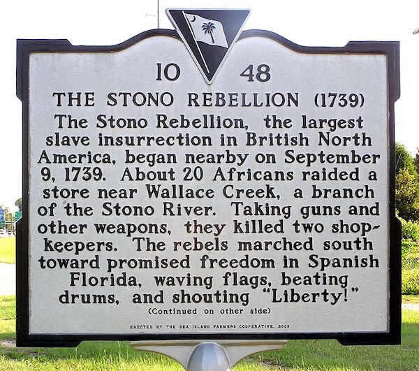 Das Foto zeigt eine Hinweistafel zur historischen Landmarke mit dem Text: "Die Stono-Rebellion Der Stono-Aufstand, der größte Sklavenaufstand in Britisch-Nordamerika, begann am 9. September 1739 in der Nähe vonbey. Etwa 20 Afrikaner überfielen einen Laden in der Nähe des Wallace Creek, einem Seitenarm des Stono River. Sie erbeuteten Gewehre und andere Waffen und töteten zwei Ladenbesitzer. Die Rebellen marschierten nach Süden in Richtung der versprochenen Freiheit im spanischen Florida, schwenkten Fahnen, schlugen Trommeln und riefen "Liberty!"