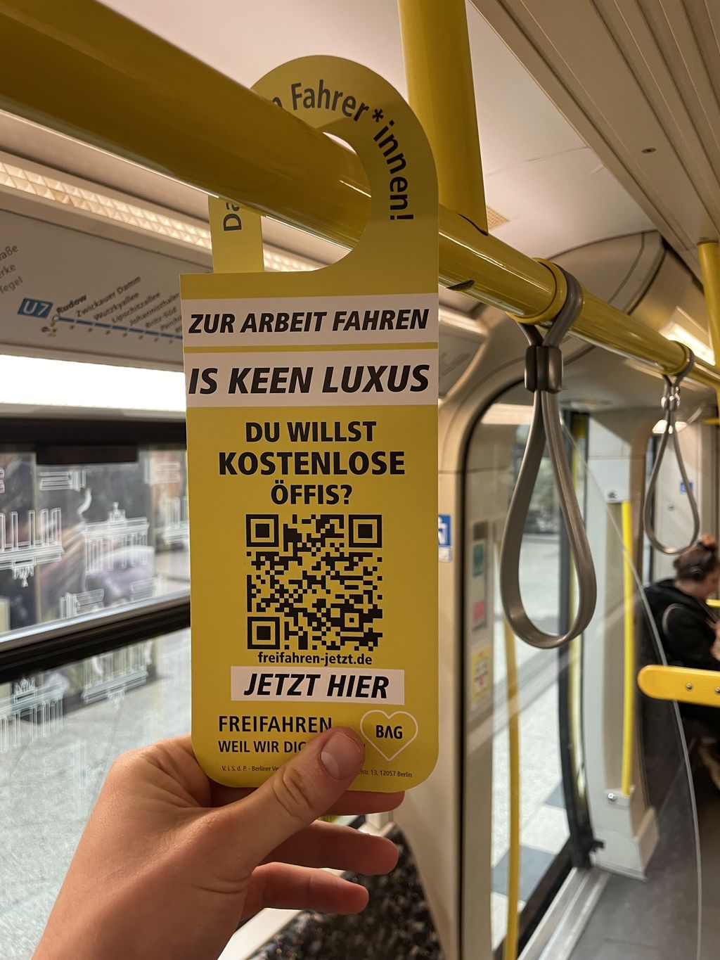 Das Foto zeigt eine Hand, die einen Hängeflyer der Aktion, der an einer Haltestange in der u-Bahn befestigt ist, hält. Auf dem Hänger sind Angaben zur Aktion sowie ein QR Code für weitere Informationen zu sehen