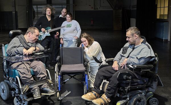 Credits: Fotografie: Werner Schmidtke Das Foto von Werner Schmidtke zeigt Darsteller:Innen des Stücks auf, neben und hinter drei Rollstühlen. Zwei Personen sitzen in je einem der Rollstühle, einer ist unbesetzt, eine Frau leht sich an den Rollstuhl