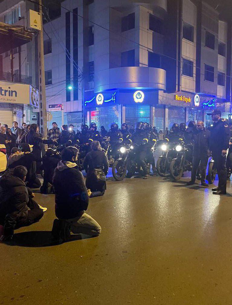 Das Foto zeigt eine abendliche Situation mit Menschen, die auf der Straße sitzen oder knien, ihnen gegenüber Sicherheitskräfte auf Motorrädern