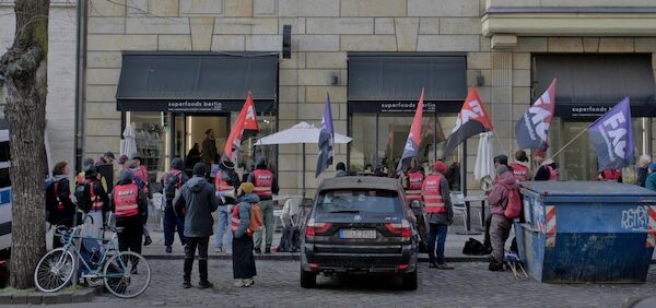 Foto: FAU Berlin Kundgebung vor dem Superfoods mit zwei Dutzend Menschen mit FAU Fahnen und Westen stehen vor dem Kundgebung vor dem Superfoods